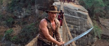 Movie still from “Indiana Jones and the Temple of Doom” (1984), directed by Steven Spielberg – A man in a hat holding a sword on top of a bridge; Medium shot, Low angle