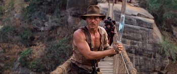 Movie still from “Indiana Jones and the Temple of Doom” (1984), directed by Steven Spielberg – A man holding a sword while standing on top of a bridge; Medium shot, Low angle