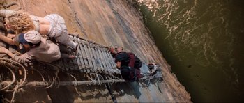 Movie still from “Indiana Jones and the Temple of Doom” (1984), directed by Steven Spielberg – A man is climbing a wooden bridge over a river; Wide shot, Overhead angle