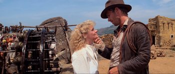 Movie still from “Indiana Jones and the Temple of Doom” (1984), directed by Steven Spielberg – A man touching a woman's face while standing on a dirt field; Medium shot, Over the shoulder angle