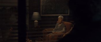 Movie still from “Infiesto” (2023), directed by Patxi Amezcua – An older woman sitting in a chair in a dark room; Wide shot, Over the shoulder angle