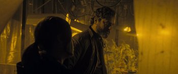 Movie still from “Infiesto” (2023), directed by Patxi Amezcua – A man standing in a dark room next to a tree; Medium shot, Over the shoulder angle