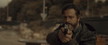 Movie still from “Infiesto” (2023), directed by Patxi Amezcua – A man holding a gun while standing in front of a brick wall; Close Up shot, Low angle