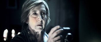 Movie still from “Insidious” (2010), directed by James Wan – An older woman looking at a camera in the dark; Close Up shot, Over the shoulder angle