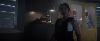 Movie still from “Interceptor” (2022), directed by Matthew Reilly – A woman standing in a dark room next to a lamp; Medium shot, Over the shoulder angle