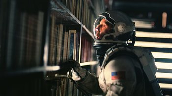 Movie still from “Interstellar” (2014), directed by Christopher Nolan – A man in a spacesuit looking at a book shelf; Medium shot, Overhead angle