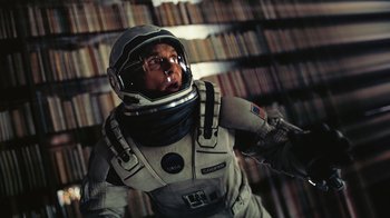 Movie still from “Interstellar” (2014), directed by Christopher Nolan – A man in a spacesuit is standing in front of a book shelf; Medium shot, Overhead angle