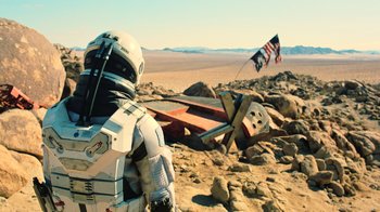Movie still from “Interstellar” (2014), directed by Christopher Nolan – A person in a spacesuit standing next to an american flag; Wide shot, Over the shoulder angle