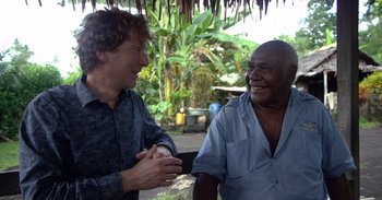 Movie still from “Into the Inferno” (2016), directed by Werner Herzog – Two men talking to each other in front of some trees; Medium shot, Over the shoulder angle