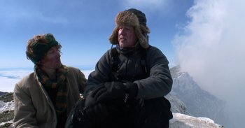 Movie still from “Into the Inferno” (2016), directed by Werner Herzog – A man wearing a winter coat and a fur hat on top of a mountain; Medium shot, Low angle