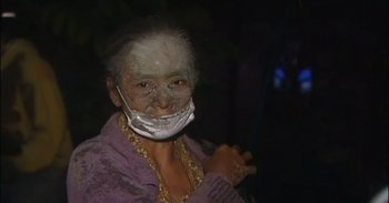 Movie still from “Into the Inferno” (2016), directed by Werner Herzog – An older woman wearing a face mask in the dark; Close Up shot, Over the shoulder angle