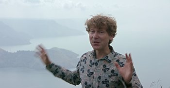 Movie still from “Into the Inferno” (2016), directed by Werner Herzog – A man standing on top of a hill with his hands up; Medium shot, Overhead angle