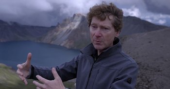 Movie still from “Into the Inferno” (2016), directed by Werner Herzog – A man standing in front of a body of water; Close Up shot, High angle