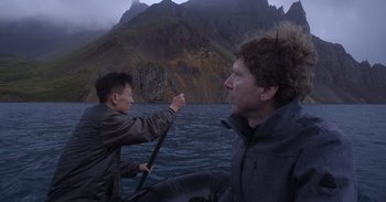 Movie still from “Into the Inferno” (2016), directed by Werner Herzog – Two men in a boat on a body of water with mountains in the background; Medium shot, High angle