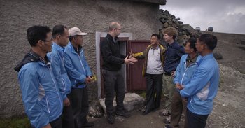 Movie still from “Into the Inferno” (2016), directed by Werner Herzog – A group of people standing around talking to each other; Medium shot, Over the shoulder angle