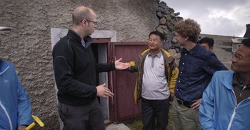 Movie still from “Into the Inferno” (2016), directed by Werner Herzog – A group of men standing next to each other near a building; Medium shot, Over the shoulder angle