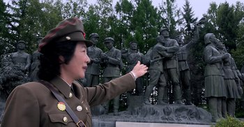 Movie still from “Into the Inferno” (2016), directed by Werner Herzog – A woman in a uniform points to a statue of a group of soldiers; Medium shot, Low angle