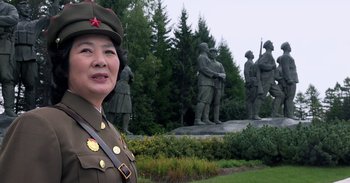 Movie still from “Into the Inferno” (2016), directed by Werner Herzog – A woman in a military uniform standing in front of statues; Medium shot, Low angle