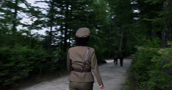 Movie still from “Into the Inferno” (2016), directed by Werner Herzog – A woman in a uniform walking down a path; Medium shot, Over the shoulder angle