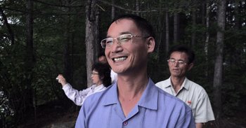 Movie still from “Into the Inferno” (2016), directed by Werner Herzog – A group of people standing in the woods with trees in the background; Medium shot, Low angle
