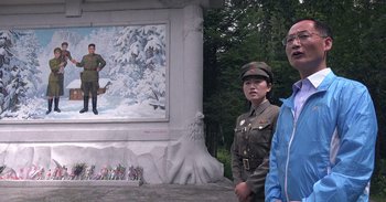 Movie still from “Into the Inferno” (2016), directed by Werner Herzog – Two people standing in front of a painting of a soldier; Wide shot, Low angle
