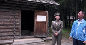 Movie still from “Into the Inferno” (2016), directed by Werner Herzog – A man in uniform standing in front of a wooden cabin; Wide shot, Low angle