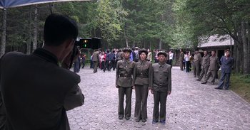 Movie still from “Into the Inferno” (2016), directed by Werner Herzog – A man taking a picture of two men in military uniforms; Wide shot, High angle