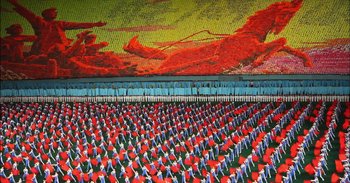 Movie still from “Into the Inferno” (2016), directed by Werner Herzog – A large group of people are standing in front of a painting; Extreme Wide shot, High angle