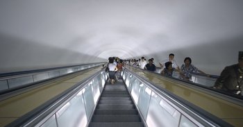Movie still from “Into the Inferno” (2016), directed by Werner Herzog – A group of people on escalators going up and down; Extreme Wide shot, High angle