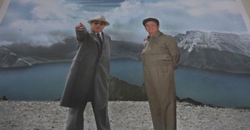 Movie still from “Into the Inferno” (2016), directed by Werner Herzog – Two men standing next to each other in front of mountains; Medium shot, Low angle