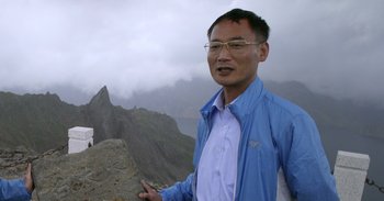 Movie still from “Into the Inferno” (2016), directed by Werner Herzog – A man standing on top of a mountain with mountains in the background; Medium shot, Low angle