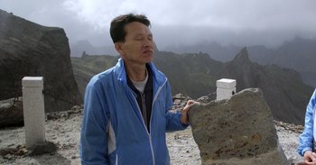 Movie still from “Into the Inferno” (2016), directed by Werner Herzog – A man standing on top of a mountain near a cliff; Medium shot, Low angle