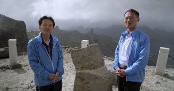 Movie still from “Into the Inferno” (2016), directed by Werner Herzog – Two people standing next to a rock on top of a mountain; Medium shot, High angle