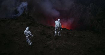 Movie still from “Into the Inferno” (2016), directed by Werner Herzog – Two people in spacesuits standing in front of an erupting volcano; Extreme Wide shot, High angle
