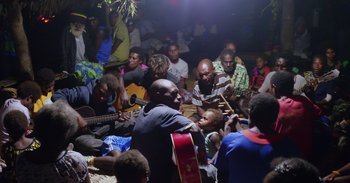 Movie still from “Into the Inferno” (2016), directed by Werner Herzog – A group of people sitting on the ground playing instruments; Medium shot, High angle