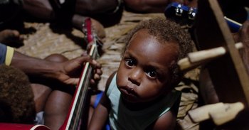 Movie still from “Into the Inferno” (2016), directed by Werner Herzog – A child is holding a guitar in his hands; Close Up shot, High angle