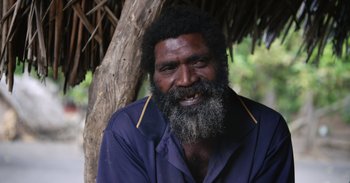 Movie still from “Into the Inferno” (2016), directed by Werner Herzog – A man with a beard and a blue shirt is smiling for the camera; Close Up shot, High angle