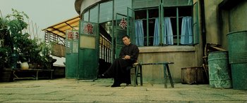 Movie still from “Ip Man 2” (2010), directed by Wilson Yip – A man sitting on top of a chair outside of a building; Wide shot, Low angle