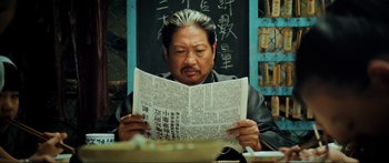 Movie still from “Ip Man 2” (2010), directed by Wilson Yip – A man sitting in front of a chalkboard holding a newspaper; Close Up shot, Low angle