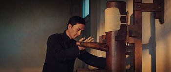 Movie still from “Ip Man 3” (2015), directed by Wilson Yip – A man in a black shirt and black shirt is doing something with a wooden stick; Medium shot, Low angle