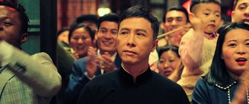 Movie still from “Ip Man 4: The Finale” (2019), directed by Wilson Yip – A group of people standing around a man in front of a wall; Close Up shot, Over the shoulder angle