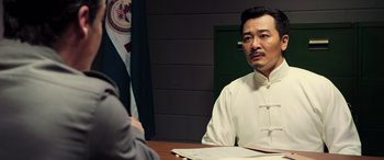 Movie still from “Ip Man 4: The Finale” (2019), directed by Wilson Yip – A man sitting at a table with papers in front of him; Medium shot, Over the shoulder angle