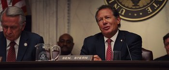 Movie still from “Iron Man 2” (2010), directed by Jon Favreau – A man sitting at a table in front of a speaker; Close Up shot, Over the shoulder angle