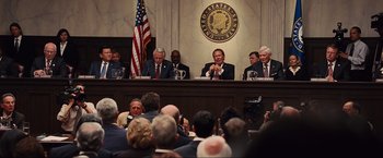 Movie still from “Iron Man 2” (2010), directed by Jon Favreau – A group of men sitting at a table in front of a flag; Wide shot, Over the shoulder angle