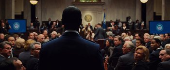 Movie still from “Iron Man 2” (2010), directed by Jon Favreau – A man in a military uniform standing in front of an audience; Medium shot, Over the shoulder angle