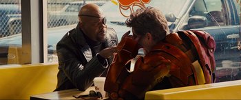 Movie still from “Iron Man 2” (2010), directed by Jon Favreau – Two men sitting at a table and one of the men is holding his hands up to his face; Medium shot, Over the shoulder angle