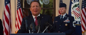 Movie still from “Iron Man 2” (2010), directed by Jon Favreau – A man in a suit and tie speaking into microphones in front of an american flag; Medium shot, Over the shoulder angle