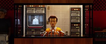 Movie still from “Isle of Dogs” (2018), directed by Wes Anderson – A television screen with a man in a orange shirt on it and microphones in front of him; Medium shot, Low angle