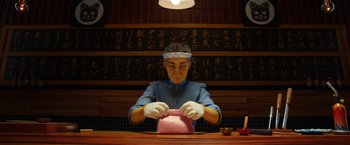 Movie still from “Isle of Dogs” (2018), directed by Wes Anderson – A person sitting at a table in front of a box; Medium shot, Low angle