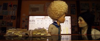 Movie still from “Isle of Dogs” (2018), directed by Wes Anderson – A doll with blonde hair sitting in front of a pile of papers; Medium shot, Low angle